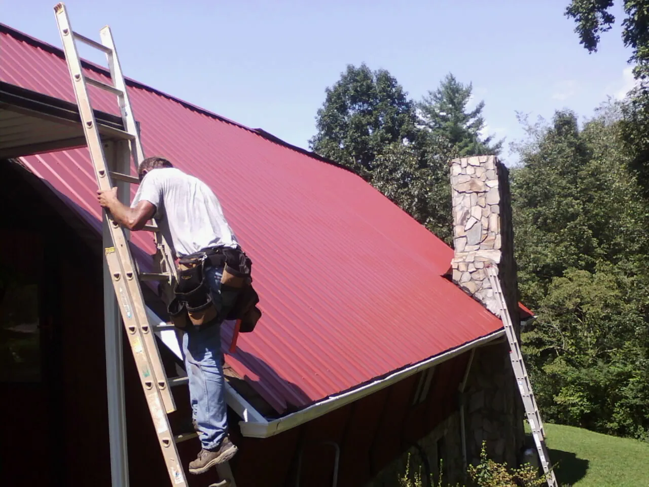 Roofer on a ladder beside a red metal roof and stone chimney — Soffit & Fascia Repair by Roofing Brothers in Loudonville